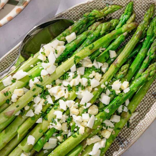 Steamed asparagus topped with a vinaigrette, grated eggs and minced chives on a platter with a serving spoon. Next to this is a cloth napkin, a stack of 2 plates, and a bowl of minced chives.