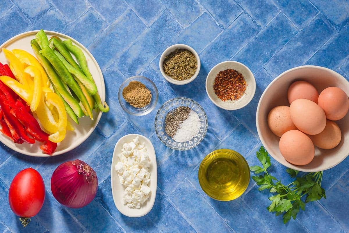 Ingredients for sheet pan eggs including eggs, bell peppers, za'atar, aleppo pepper, cumin, red onion, salt, black pepper, olive oil, parsley, feta and a tomato.