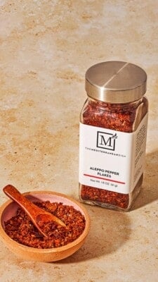 A bottle of Aleppo pepper from The Mediterranean Dish shop next to a small bowl of the pepper with a spoon.