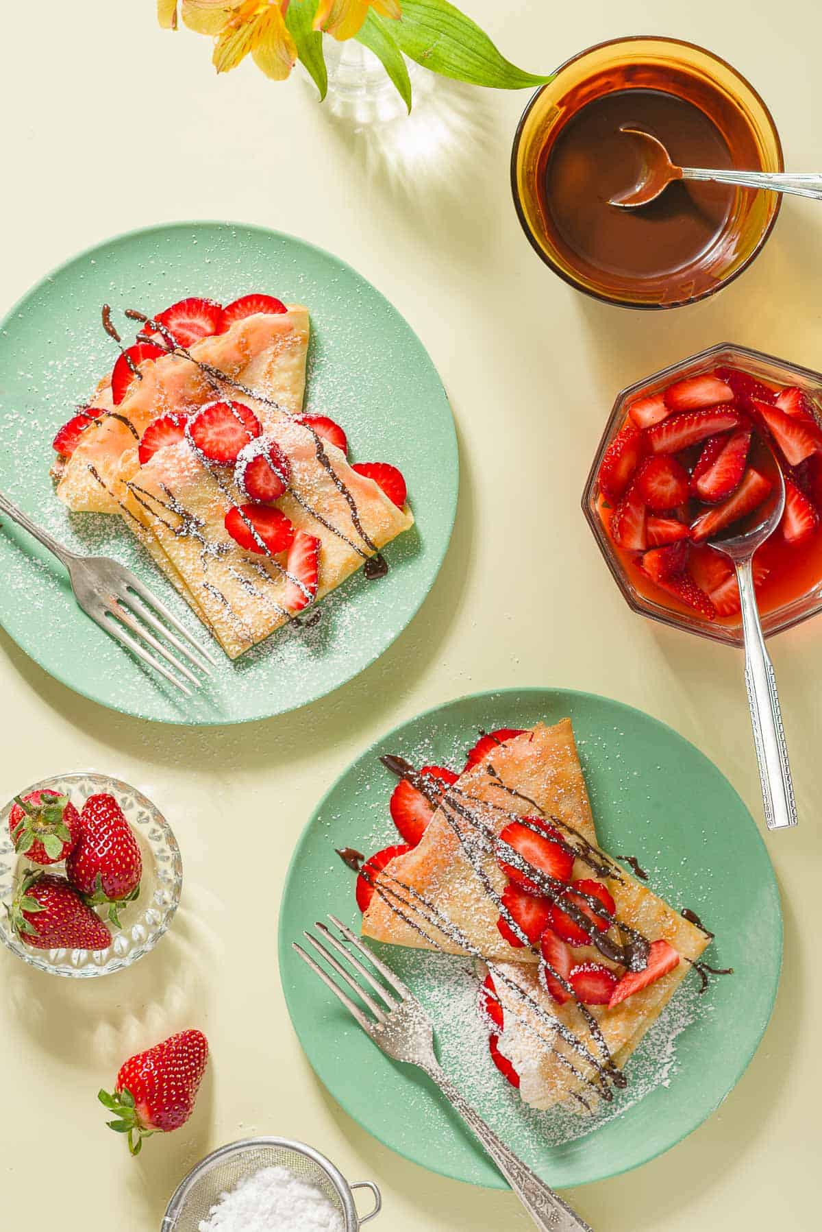 Two plates, each containing 3 strawberry crepes and a fork, surrounded by powdered sugar in sifter, whole strawberries in a bowl, a bowl of macerated strawberries with a spoon, and a bowl of melted chocolate with a spoon.