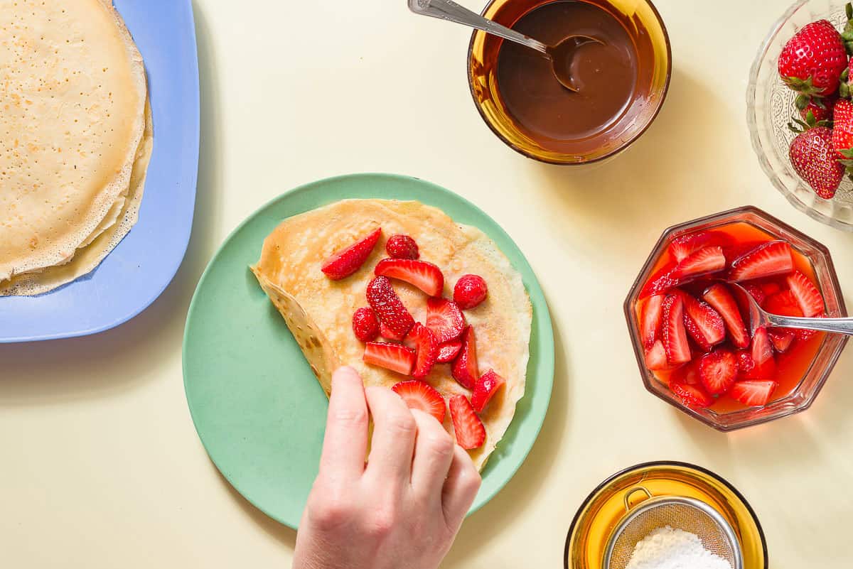 A crepe on a plate being topped with strawberries. Next to this is a stack of crepes on a platter, a bowl of powdered sugar, a bowl of melted chocolate with a spoon, a bowl of macerated strawberries with a spoon, and a bowl of whole strawberries.
