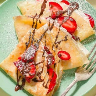 Three strawberry crepes drizzled with chocolate on a plate with a fork.