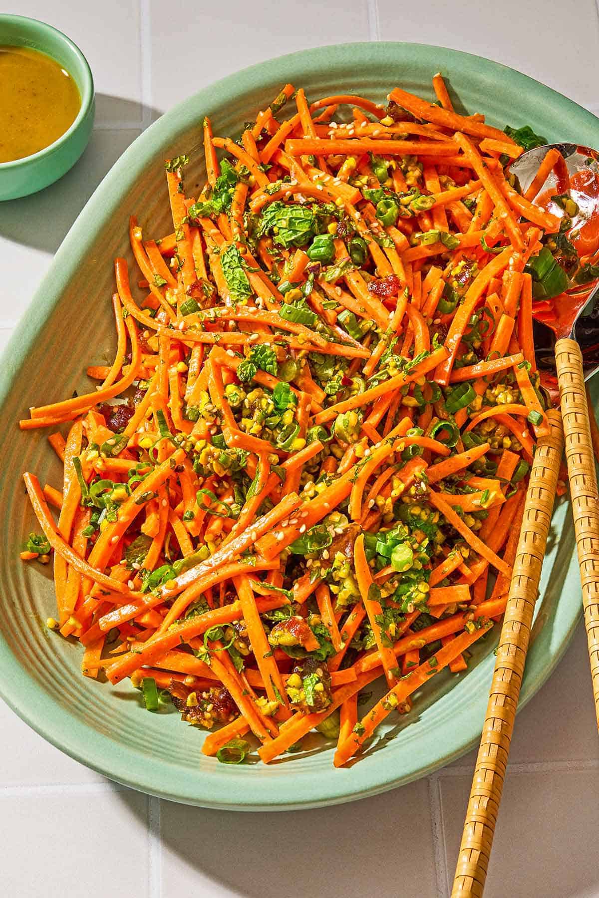 Carrot salad with herbs on a platter with serving utensils next to a bowl of the dressing.