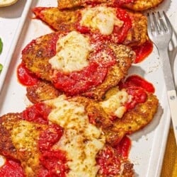Six pieces of chicken parmesan on a platter with a fork. Next to this is a bowl of grated Parmigiano-Reggiano cheese.