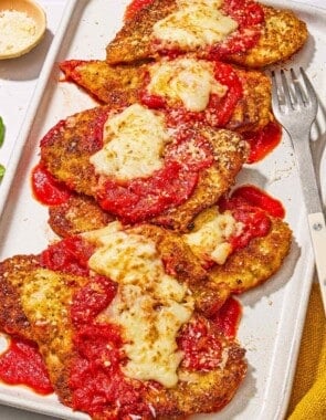 Six pieces of chicken parmesan on a platter with a fork. Next to this is a bowl of grated Parmigiano-Reggiano cheese.