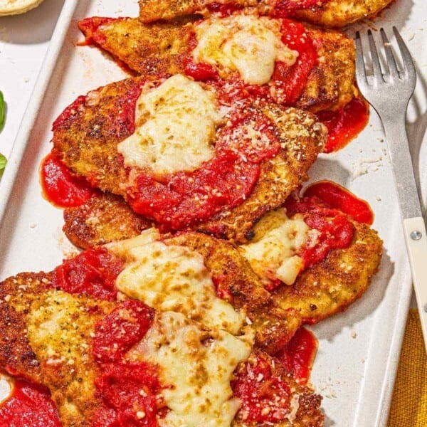 Six pieces of chicken parmesan on a platter with a fork. Next to this is a bowl of grated Parmigiano-Reggiano cheese.