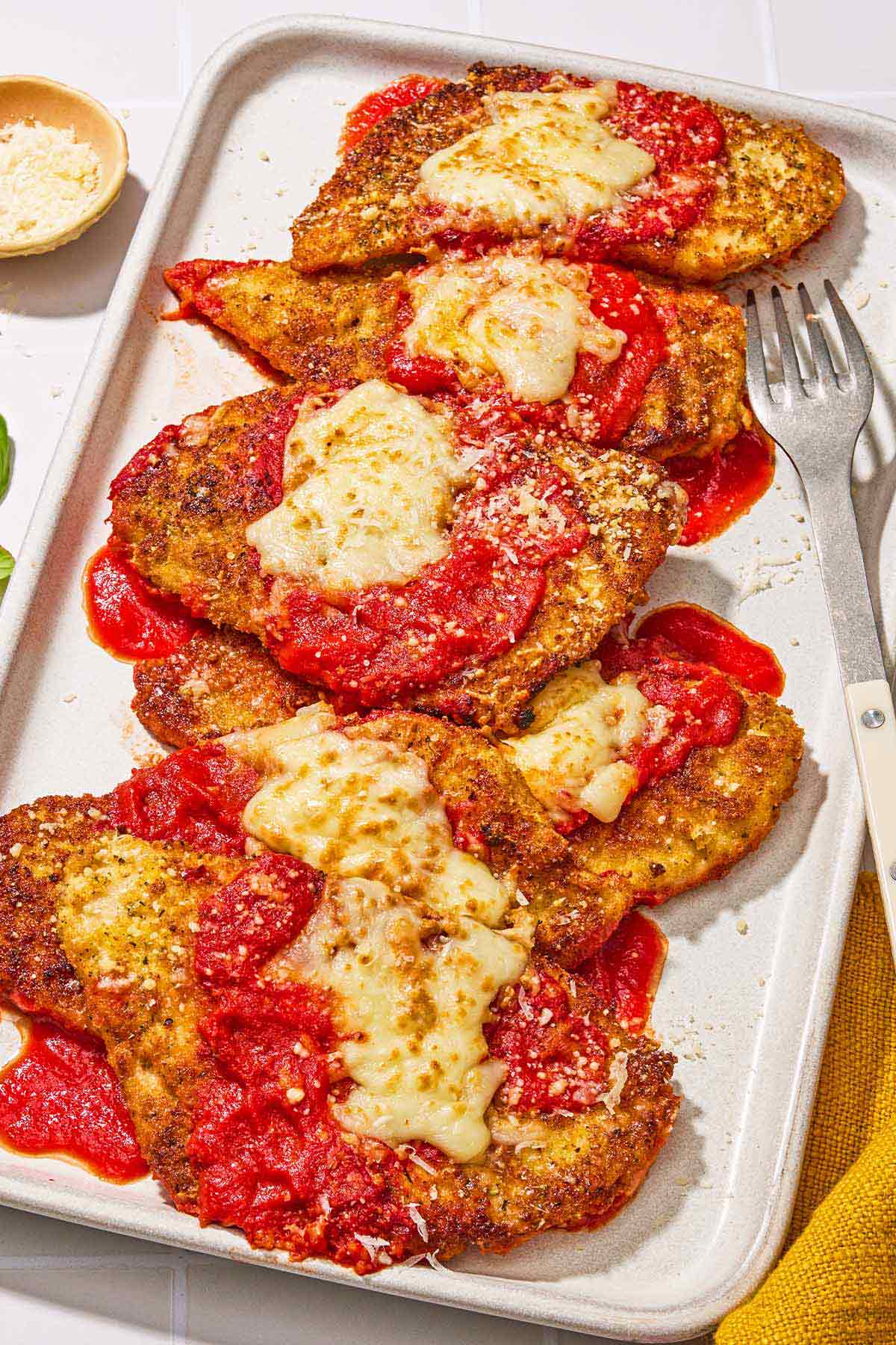 Six pieces of chicken parmesan on a platter with a fork. Next to this is a bowl of grated Parmigiano-Reggiano cheese.