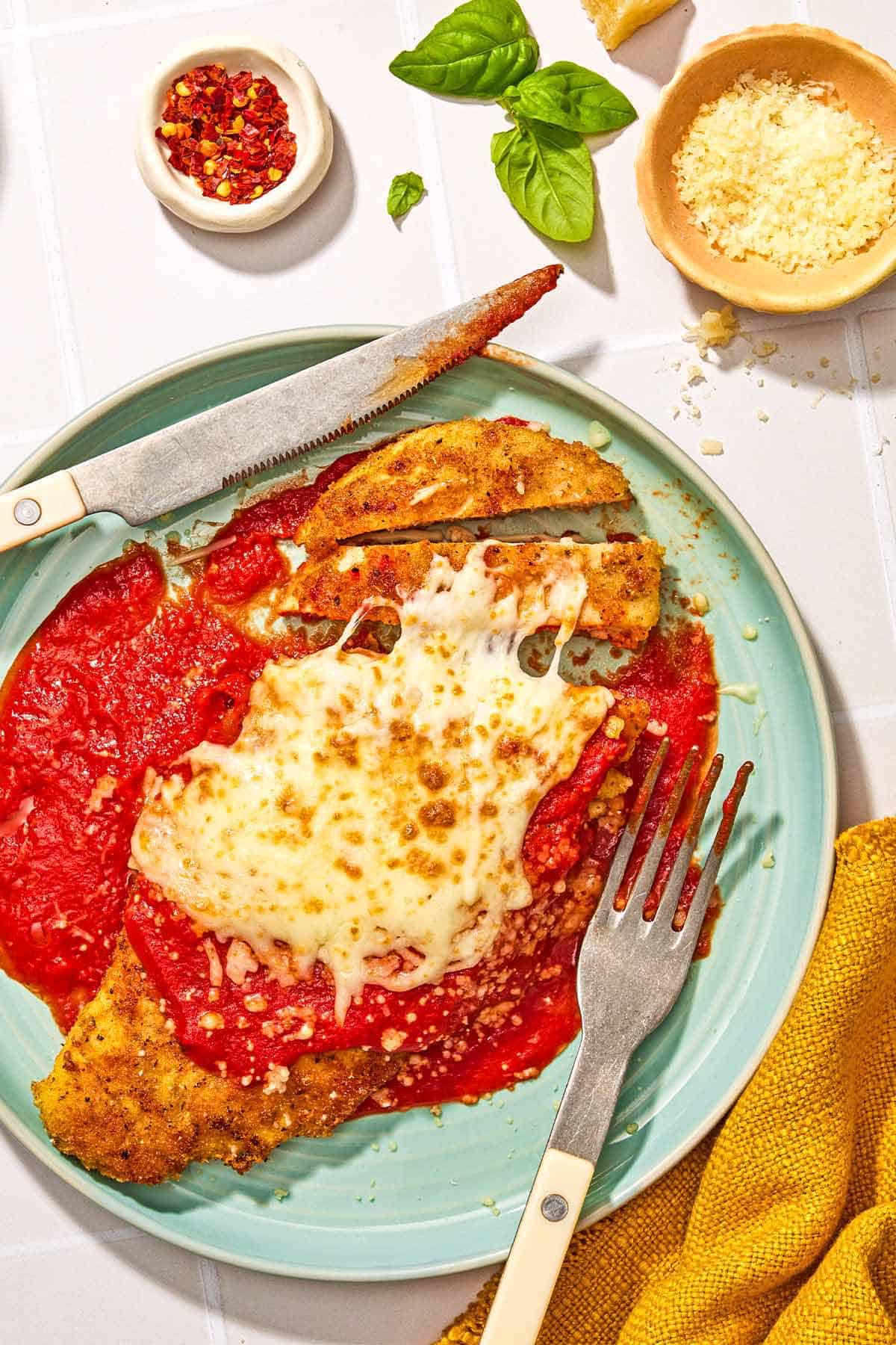 One piece of the chicken parmesan on a plate with a knife and fork. Next to this is a bowl of red pepper flakes, a few basil leaves, a bowl of grated Parmigiano-Reggiano cheese and a cloth napkin.