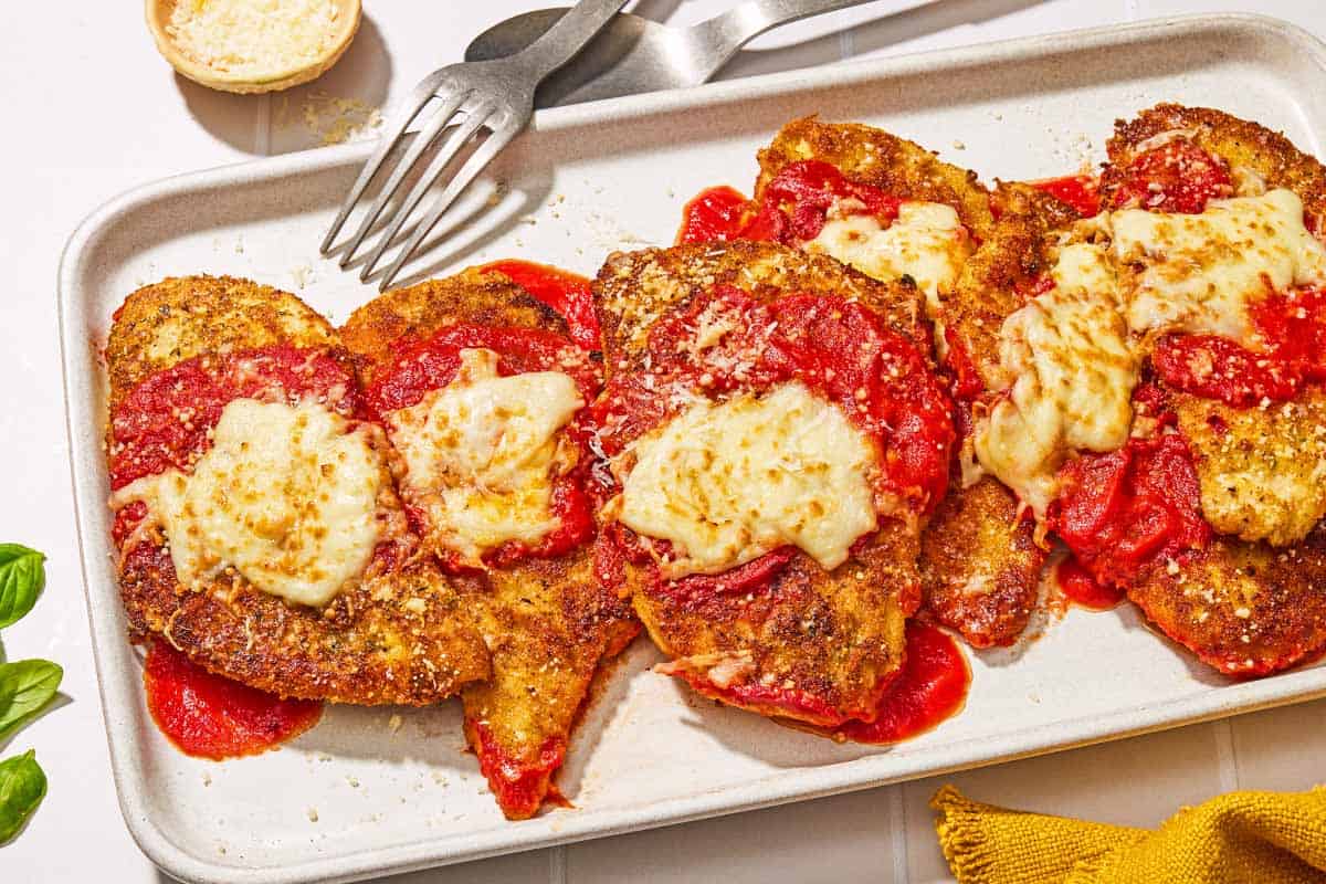 Six pieces of chicken parmesan on a platter with a fork. Next to this is a bowl of grated Parmigiano-Reggiano cheese and a serving spoon.