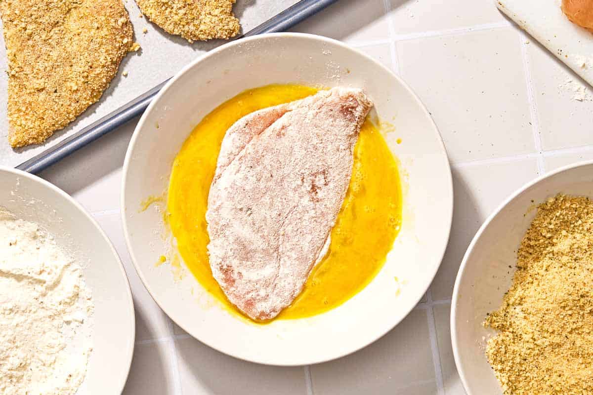 A floured chicken cutlet for the chicken parmesan recipe in a bowl with lightly beaten egg. next to this is a bowl of flour, a bowl of breadcrumbs and a baking sheet with other breaded but uncooked chicken cutlets.