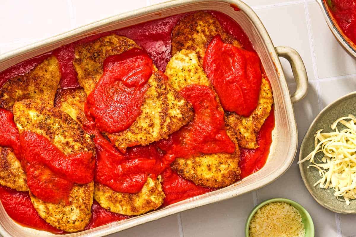 The cooked, breaded chicken cutlets for the chicken parmesan in a baking dish with the sauce. Next to this is a bowl of grated Parmigiano-Reggiano cheese and a bowl of shredded mozzarella.