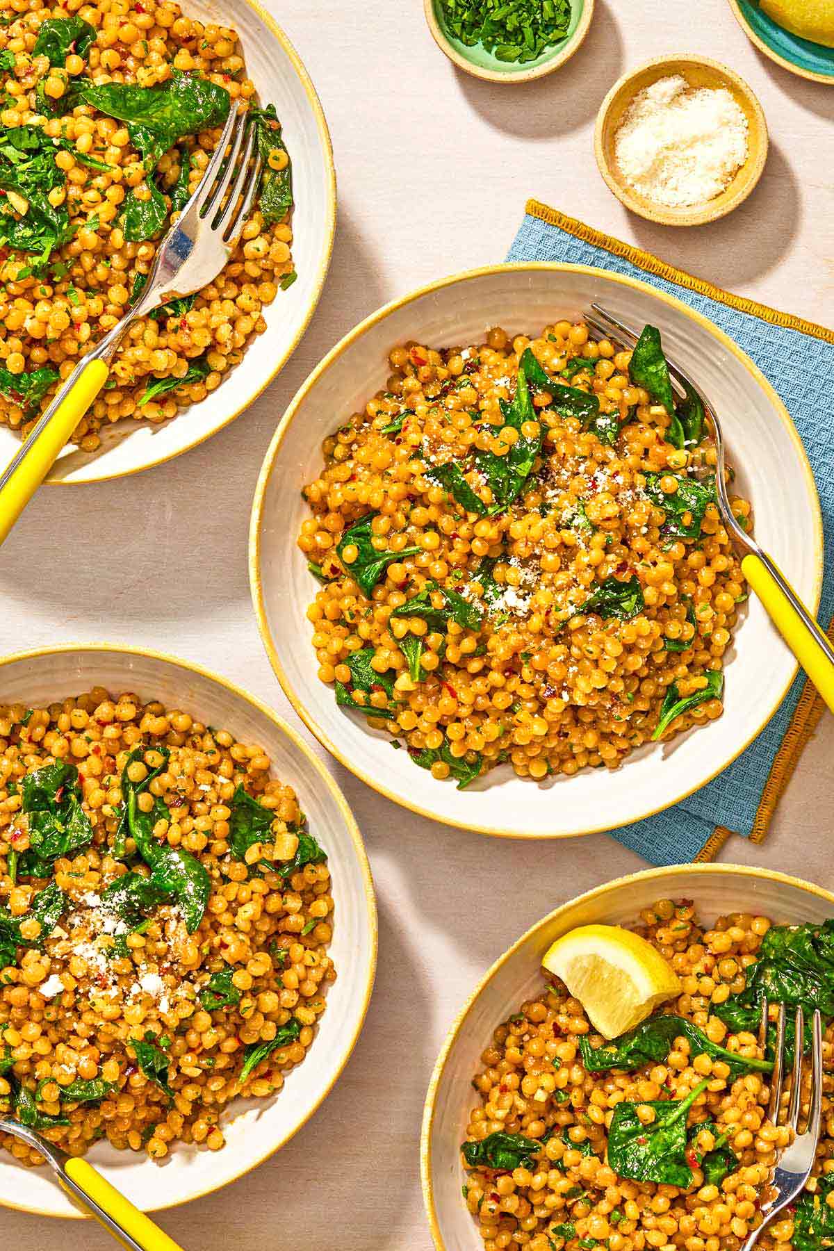 Four bowls of spinach couscous with forks. Next to these are bowls of grated Pecorino Romano cheese and chopped parsley, a cloth napkin, and a plate of lemon wedges.