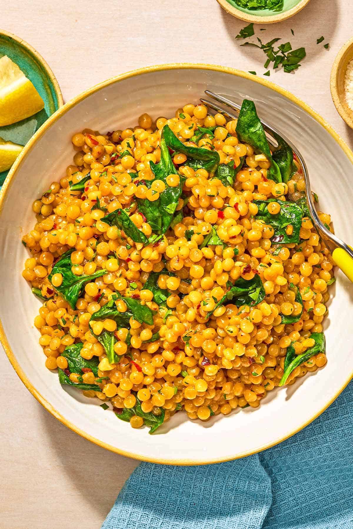 Spinach couscous in a bowl with a fork. Next to this is a cloth napkin, a plate of lemon wedges, and bowls of grated Pecorino Romano cheese and chopped parsley.