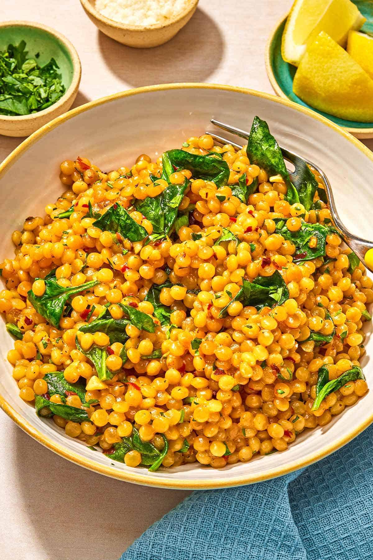 Spinach couscous in a bowl with a fork. Next to this is a cloth napkin, a plate of lemon wedges, and bowls of grated Pecorino Romano cheese and chopped parsley.