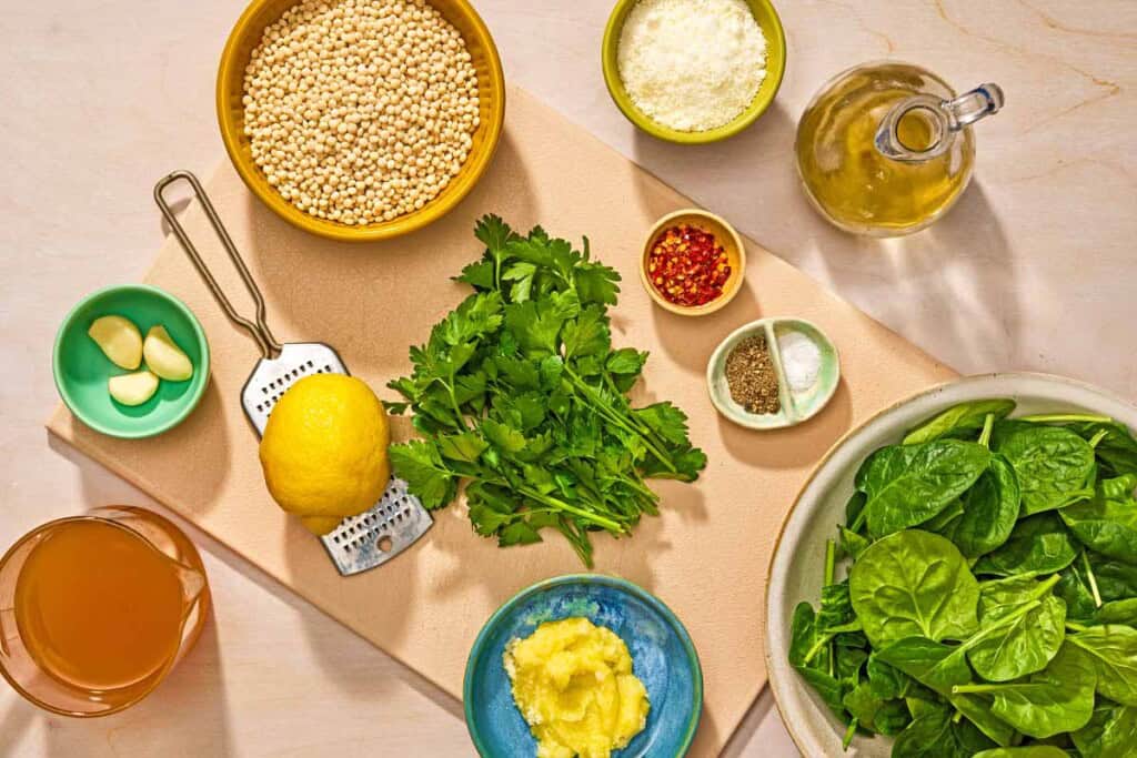 Ingredients for spinach couscous including pearl couscous, fresh spinach, garlic, red pepper flakes, olive oil, garlic, broth, ghee, Pecorino Romano cheese, parsley and lemon.