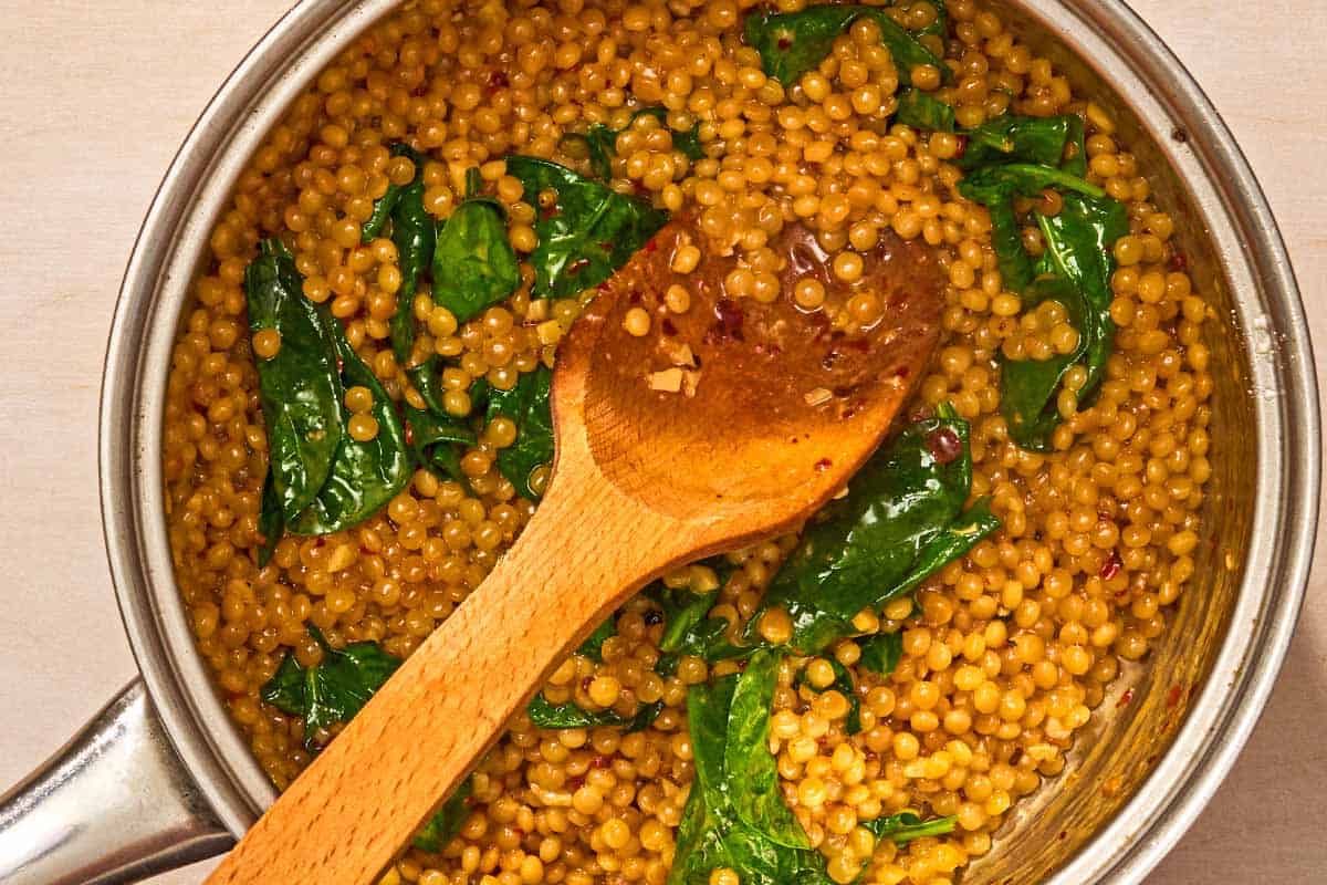 The baby spinach, ghee, and Pecorino Romano cheese being stirred into the cooked couscous in a saucepan with a wooden spoon.