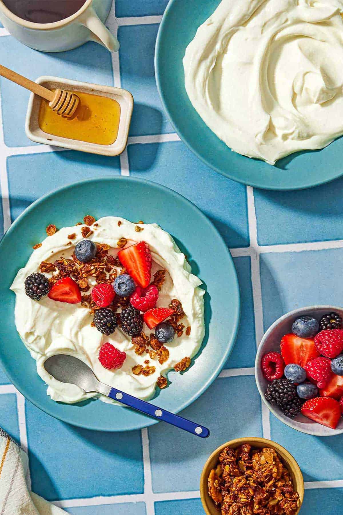 A bowl of homemade greek yogurt topped with a mix of berries and granola in a bowl with a spoon surrounded by bowls of honey, berries and granola, and another bowl of plain homemade greek yogurt.