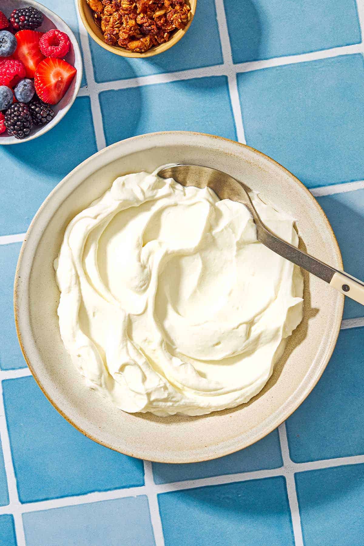 Homemade greek yogurt in a bowl with a spoon next to a small bowl with various berries and a bowl of granola.