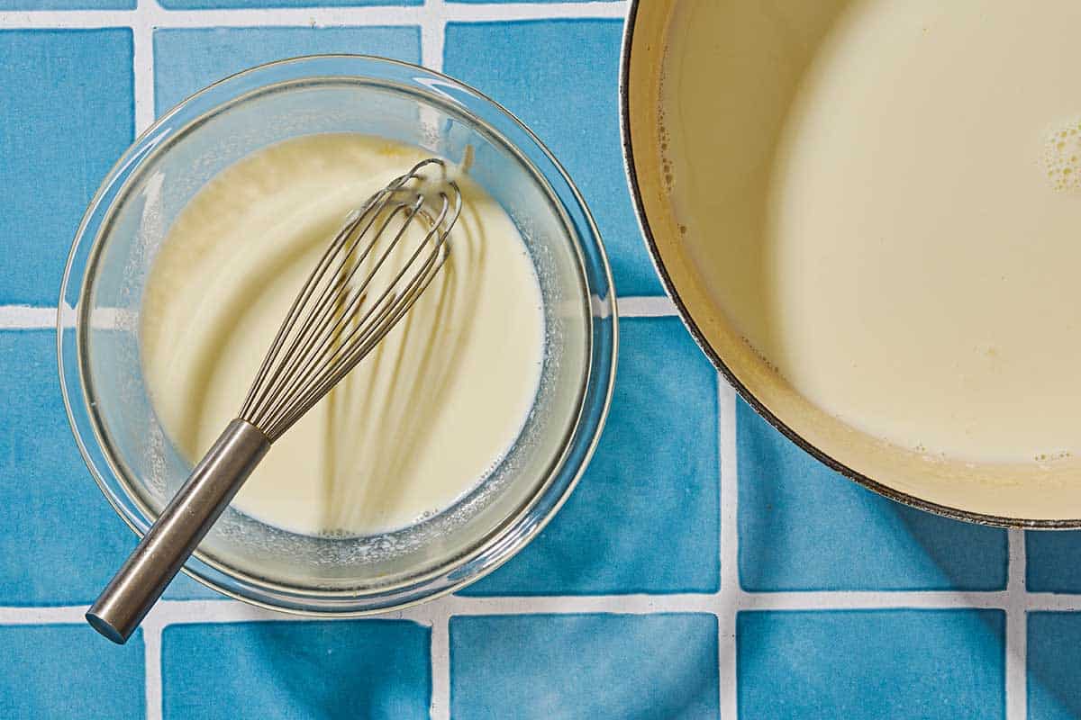 Warm milk in a pot next to a heat safe bowl containing the starter yogurt mixed with a bit of the milk, along with a whisk.
