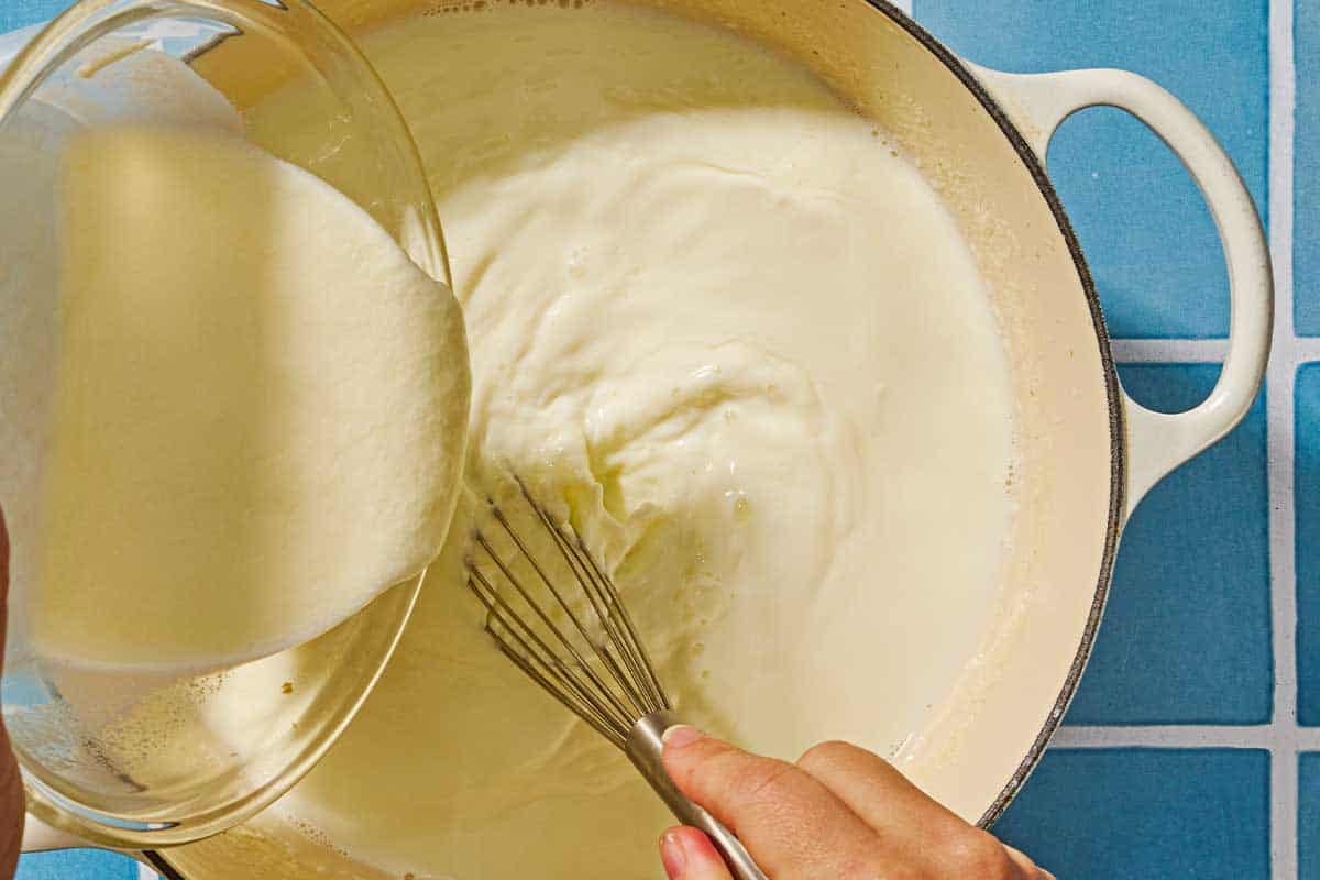 The milk and stater yogurt mixture being poured into the pot with the warm milk as it's being stirred with a whisk.