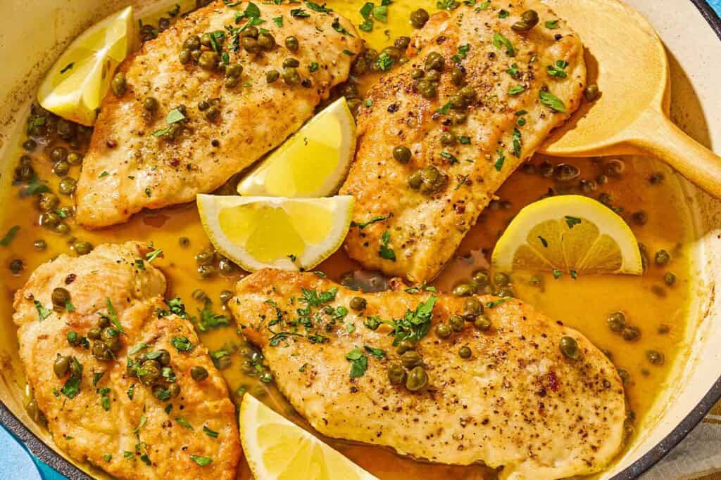 Chicken piccata with lemon wedges in a skillet with a wooden spoon.