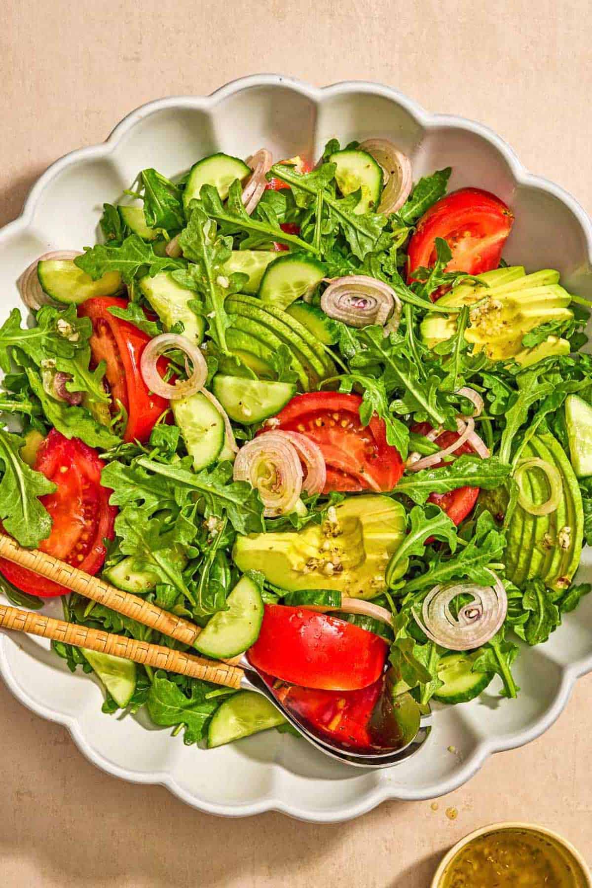 Rocket salad with tomato, cucumber and avocado in a bowl with serving tools next to a bowl of the sauce.