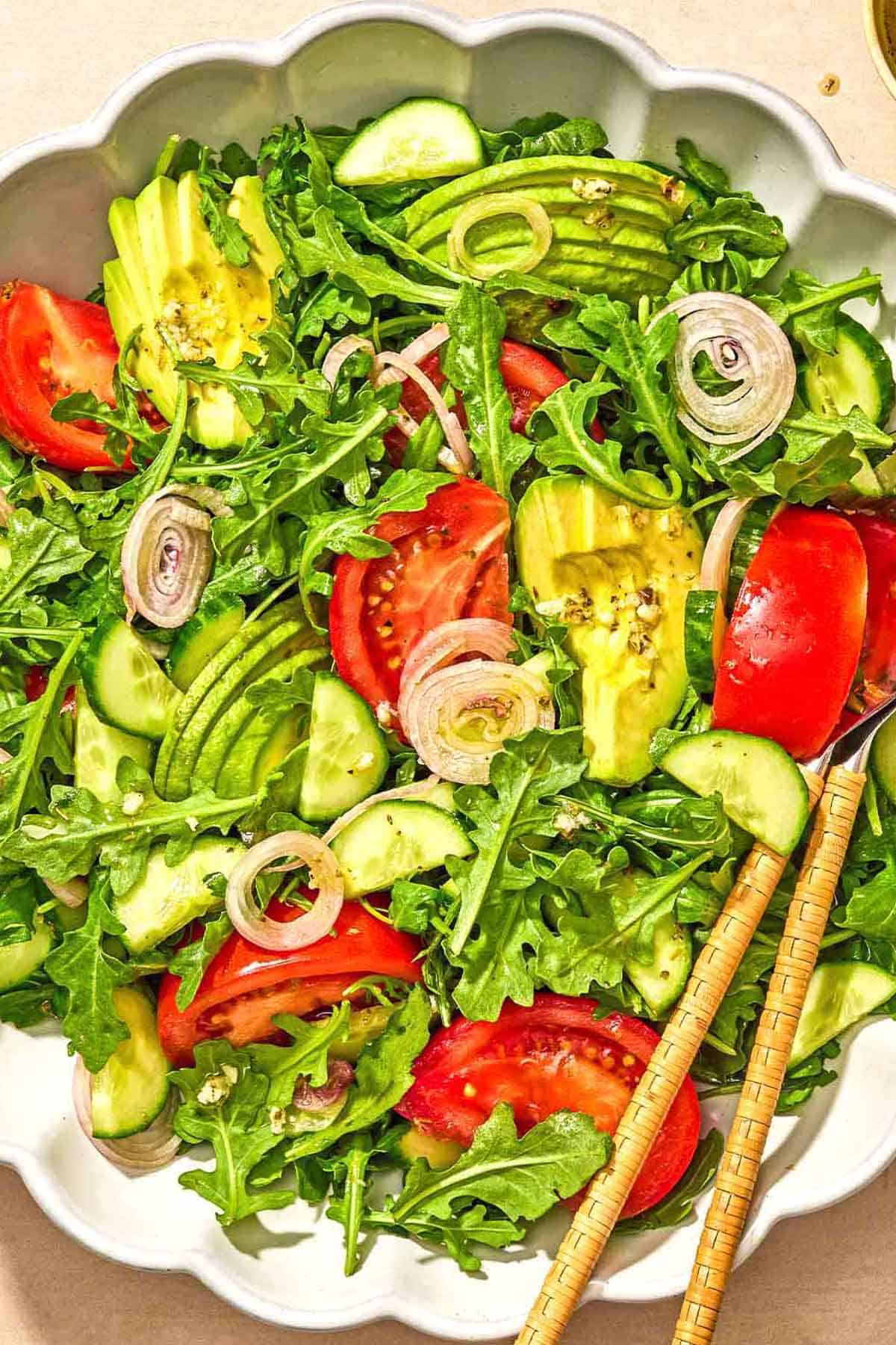 Rocket salad with tomato, cucumber and avocado in a bowl with serving utensils.