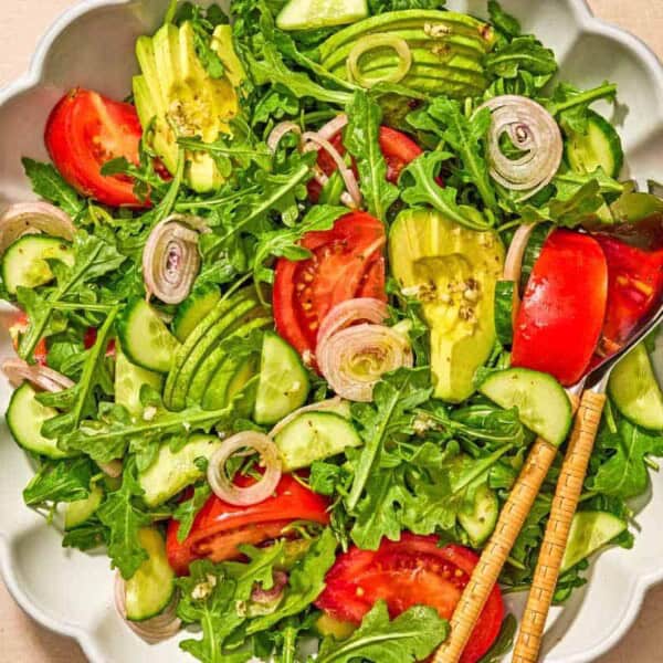 Arugula salad with tomato, cucumber and avocado in a serving bowl with serving utensils next to a bowl of the dressing.