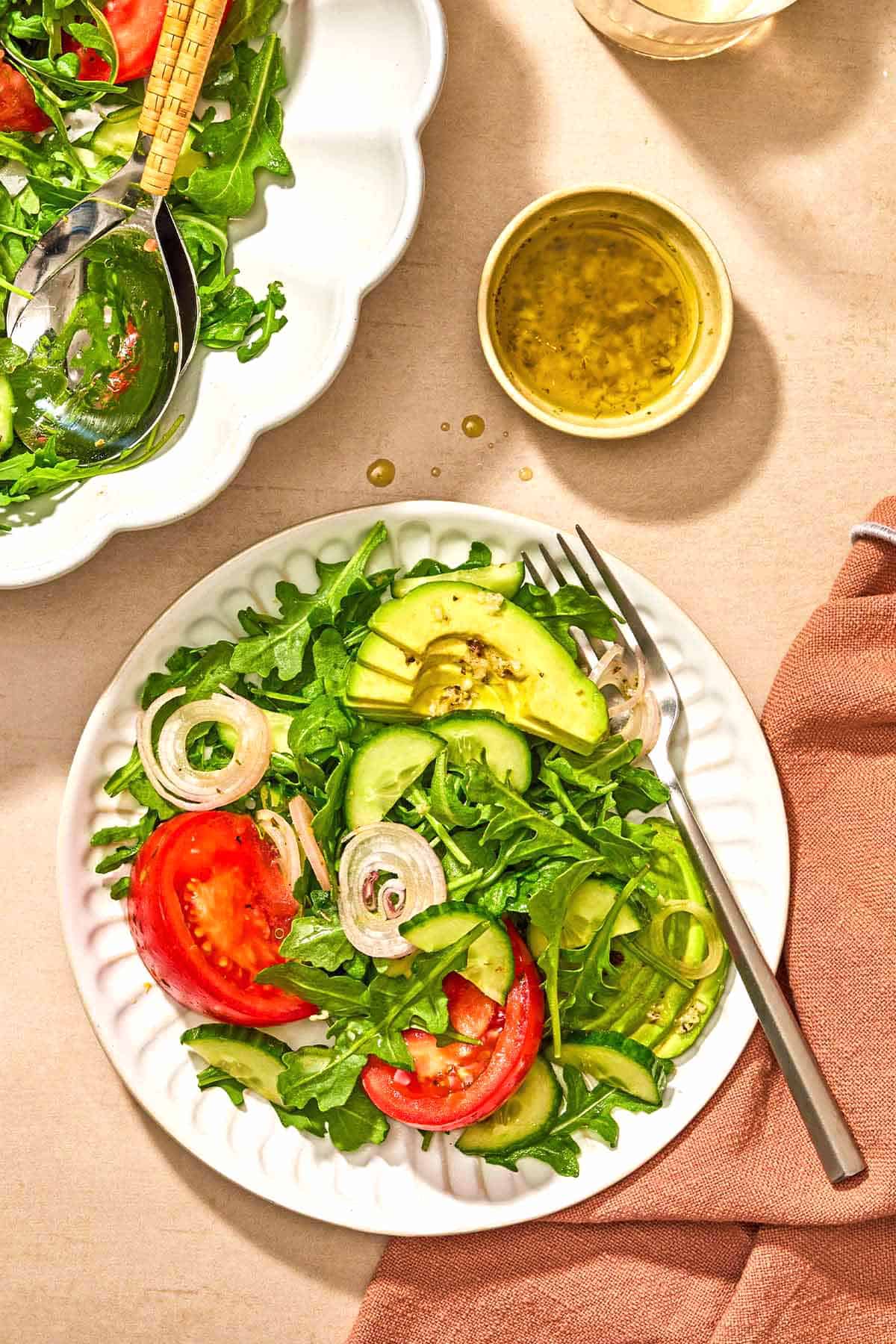Rocket salad with tomato, cucumber and avocado on a plate with fork. Next to this there is a fabric napkin, a small bowl with salad dressing and the rest of the exhausted in a serving bowl with serving tools.