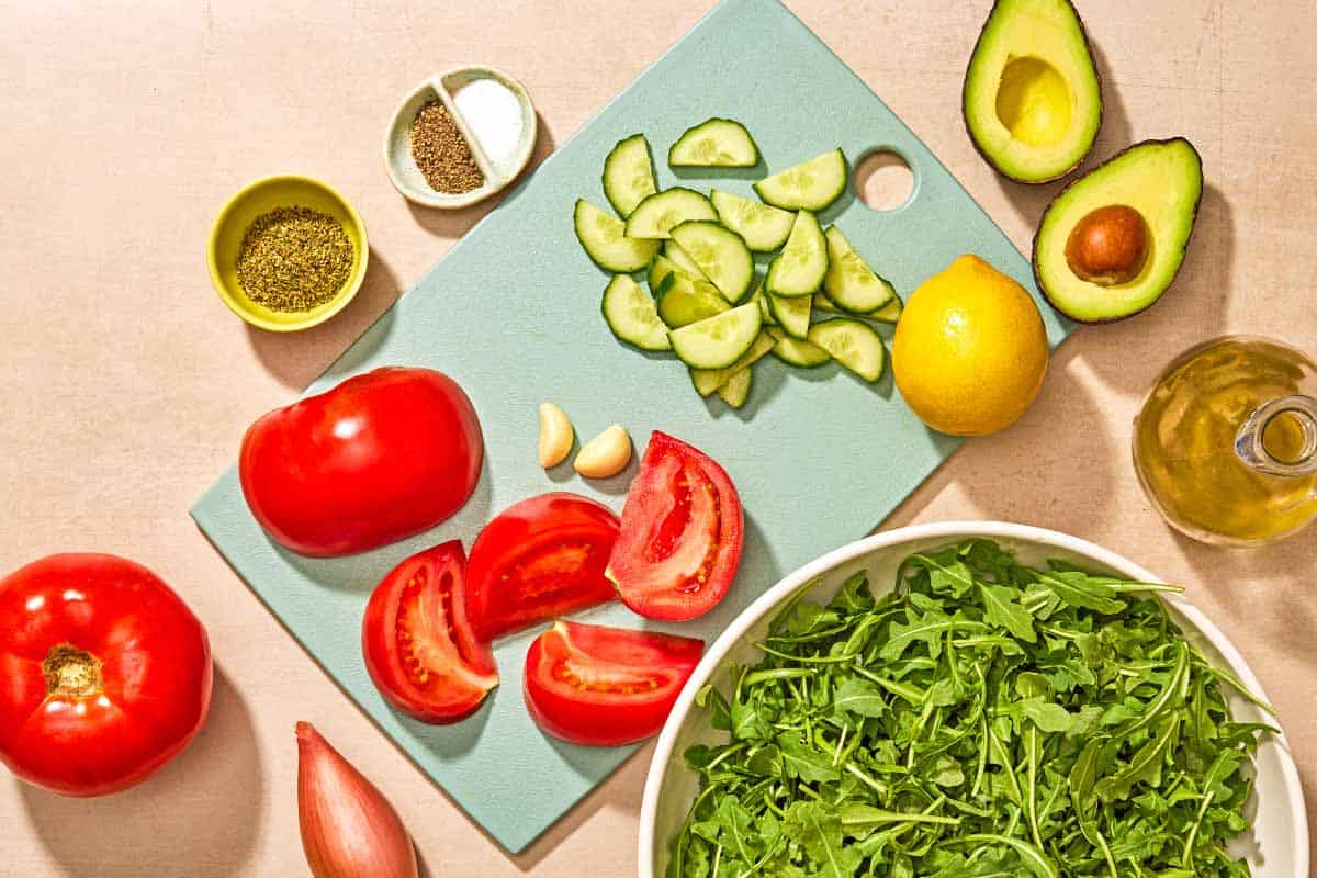 Ingredients for rocket salad including rocket, tomato, cucumber, avocado, lemon, garlic, a shallot, olive oil, salt, black pepper and dry oregano.