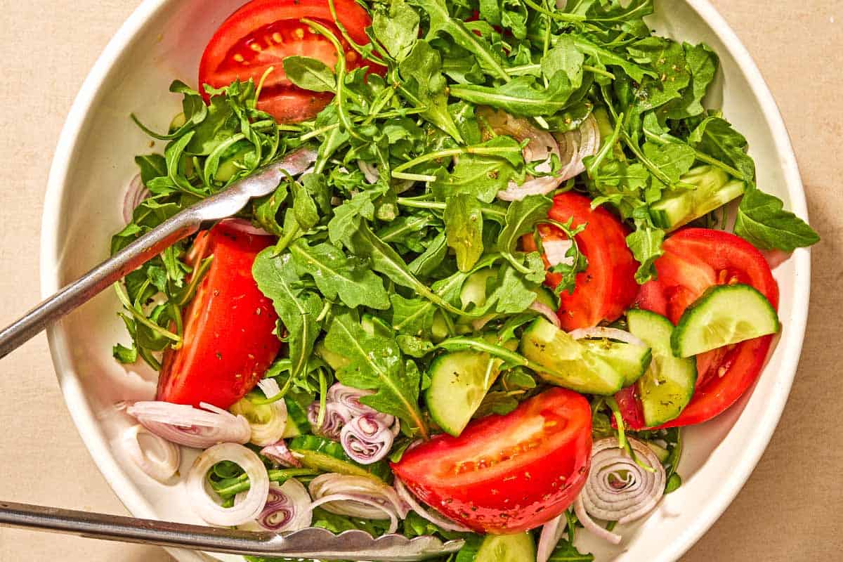 Arugula salad with tomato, cucumber and avocado in a serving bowl with tongs.