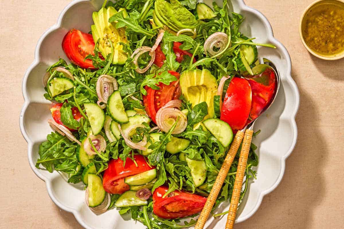 Arugula salad with tomato, cucumber and avocado in a serving bowl with serving utensils next to a bowl of the dressing.