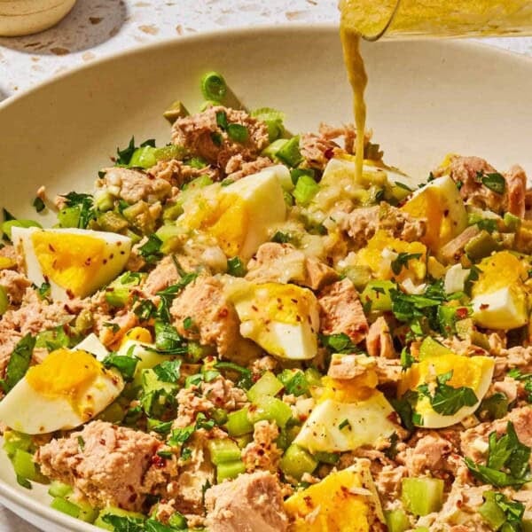 The dijon vinaigrette being poured onto the tuna salad with egg in a serving bowl with a small bowl of aleppo pepper in the background.