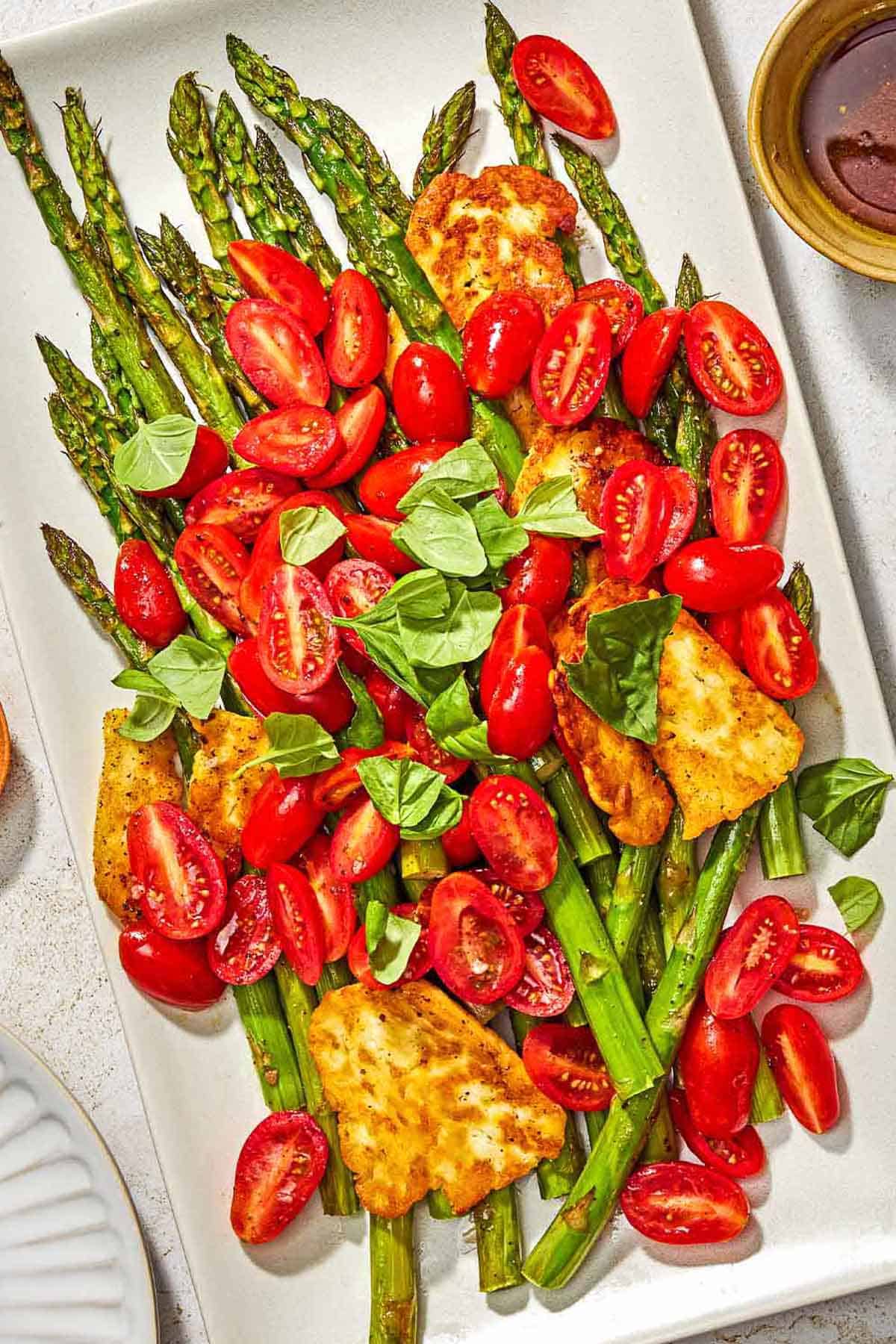 Roasted Asparagus Salad with Fried Halloumi and Fresh Tomatoes on a serving platter next a bowl of the sherry vinaigrette.