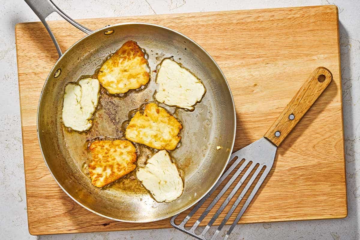 Six slices of fried halloumi cheese in a skillet next to a spatula on a wooden serving board.