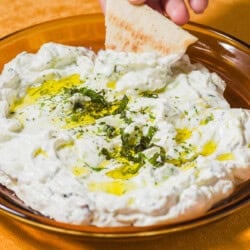 A pita wedge being dipped into a bowl of Maast O'Khiar garnished with dried mint and a drizzle of olive oil.