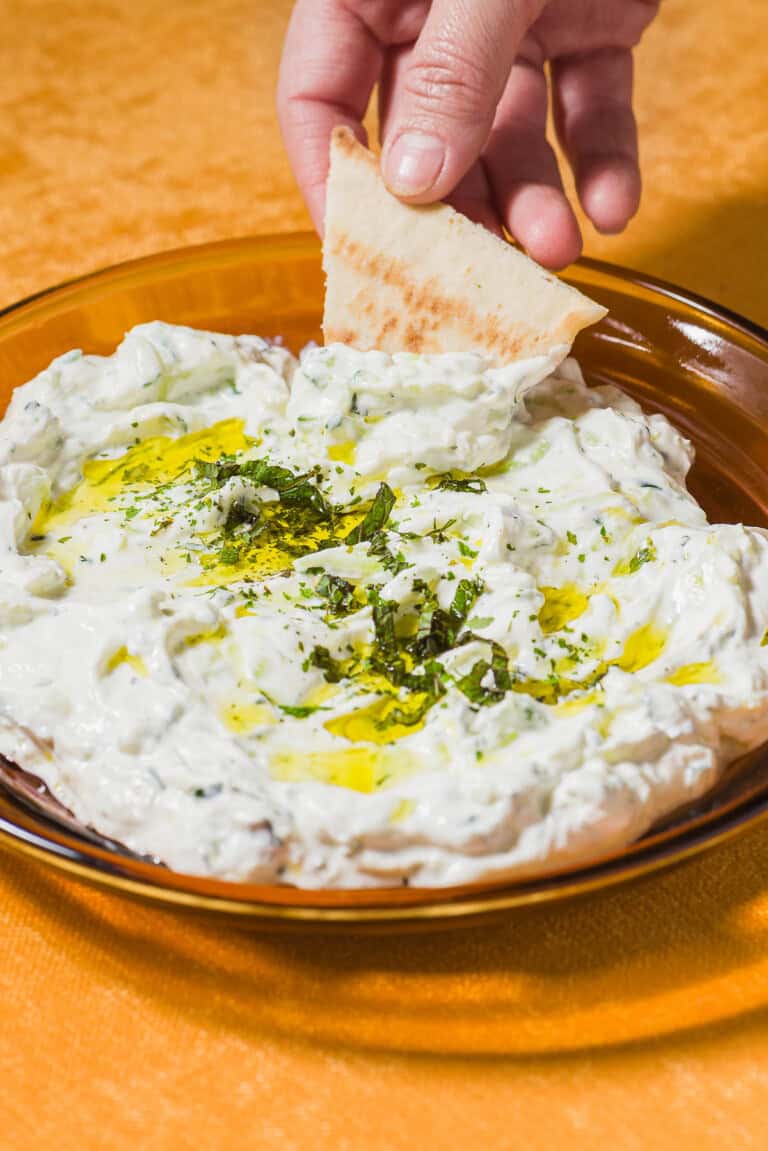 A pita wedge being dipped into a bowl of Maast O'Khiar garnished with dried mint and a drizzle of olive oil.