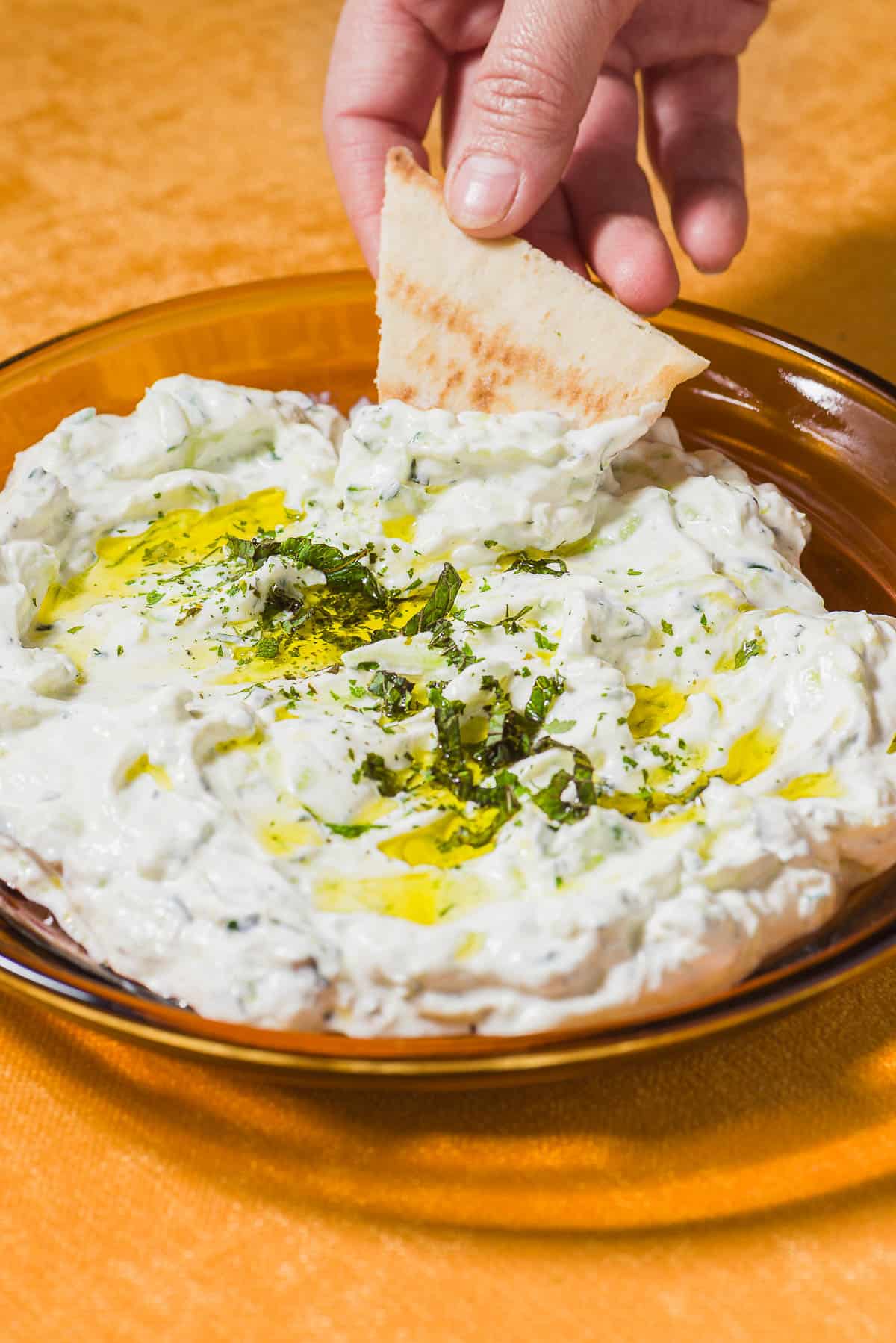 A pita wedge being dipped into a bowl of Maast O'Khiar garnished with dried mint and a drizzle of olive oil.