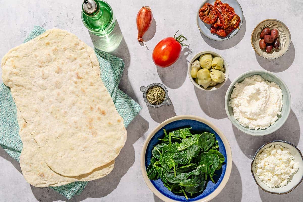 Ingredients for flatbread pizza including flatbread, olive oil, ricotta cheese, feta, oregano, marinated artichoke hearts, baby spinach, tomato, shallot, kalamata olives and sun-dried tomatoes.