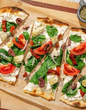 Slices of the flatbread pizza on parchment paper surrounded by bowls of kalamata olives, oregano and sliced tomato.
