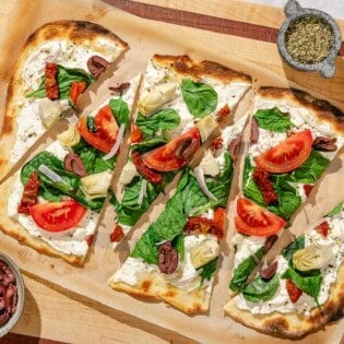 Slices of the flatbread pizza on parchment paper surrounded by bowls of kalamata olives, oregano and sliced tomato.