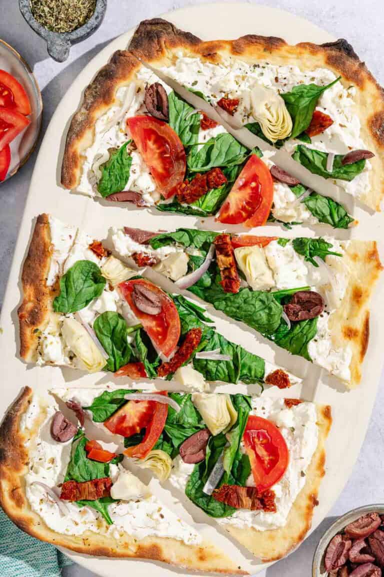 Slices of the flatbread pizza on a platter surrounded by bowls of kalamata olives, oregano and sliced tomato.