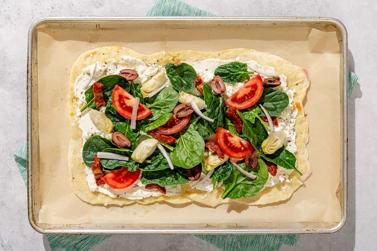 The unbaked flatbread topped with ricotta cheese, feta cheese, oregano, baby spinach, tomato, shallot, kalamata olives, sun dried tomatoes and artichoke hearts on a parchment paper lined sheet pan.