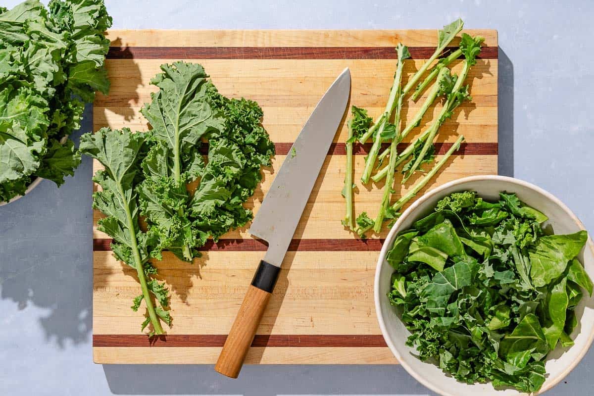 A cutting board with whole kale leaves, a knife, kale stems and a bowl with the trimmed greens.