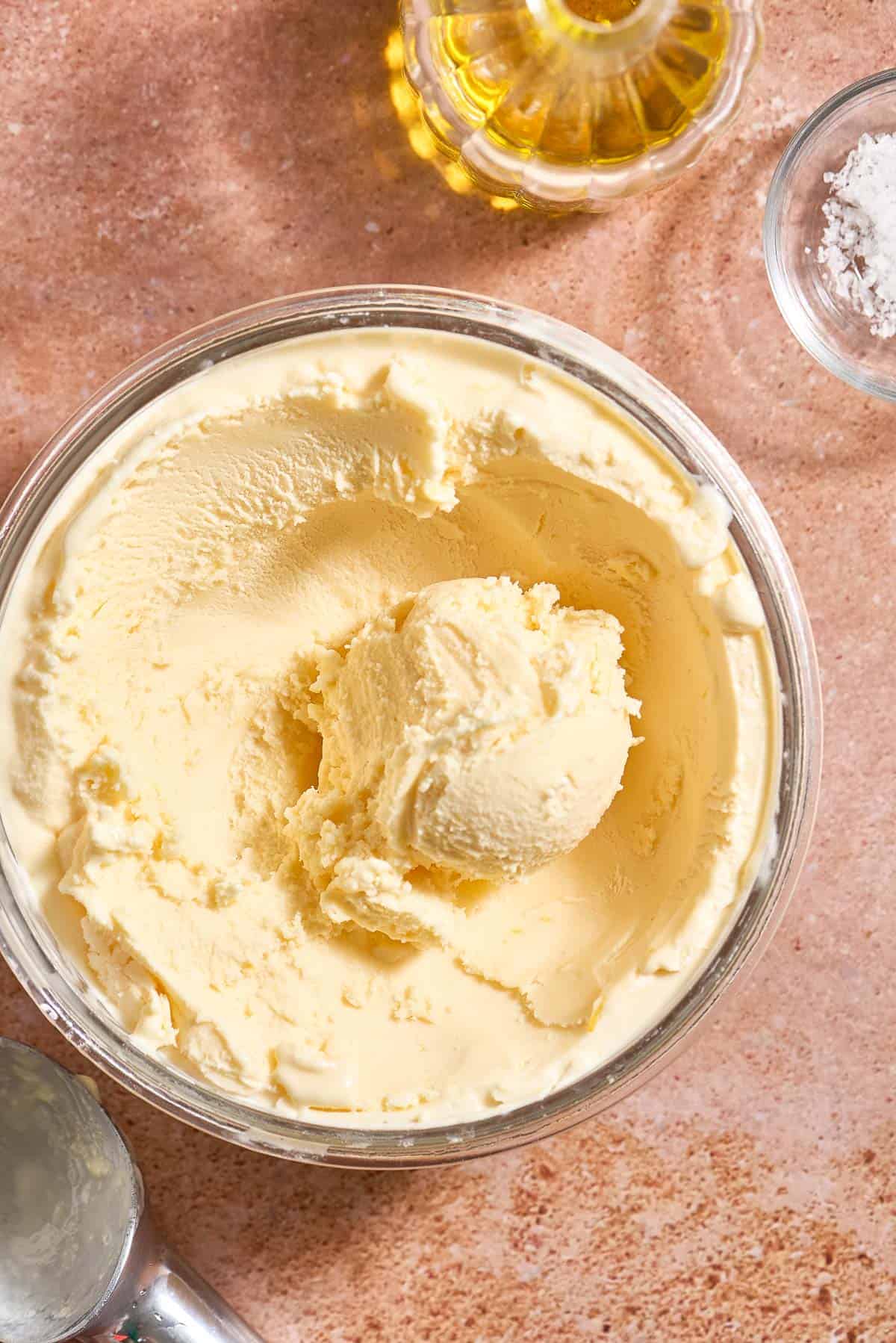 Olive oil ice cream in a large glass container next to a bottle of olive oil, a bowl of flaky sea salt and an ice cream scoop.
