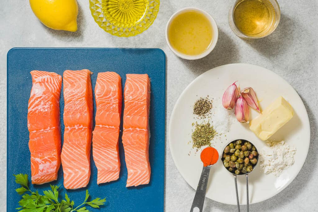 The ingredients for salmon piccata including salmon, sweet paprika, oregano, salt, black pepper, olive oil, butter, garlic, flour, chicken broth, white wine, lemon, capers, parsley and lemon.