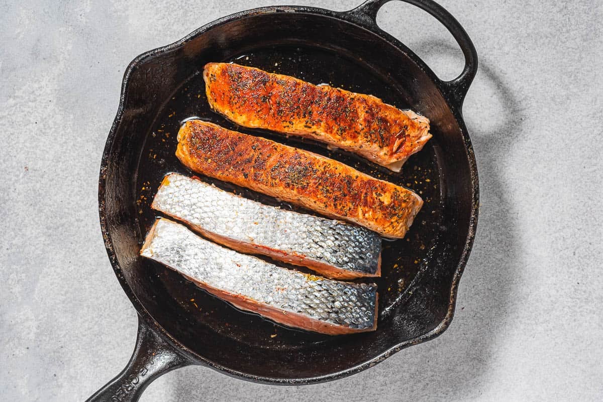 4 salmon fillets being seared in a cast iron skillet. Two are skin up and 2 are flesh up.