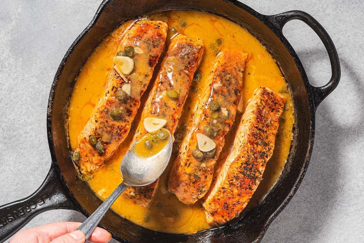 Sauce being spooned over the 4 salmon piccata fillets in a cast iron skillet.