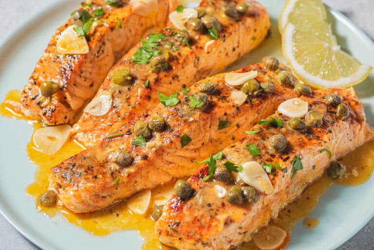 4 salmon piccata fillets on a plate with lemon wedges.