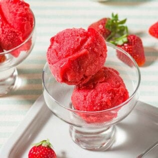 Two scoops of strawberry sorbet in a bowl next to a spoon, several scattered strawberries, a small bowl of strawberries and another bowl of the sorbet.